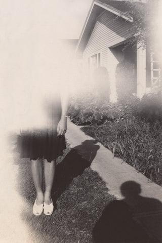 Black and white vintage photograph of a young woman with a blurred camera effect covering her face, with a silhouette of the photographer in the foreground 